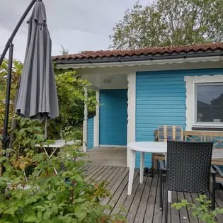 Trädäck med bord och stolar framför en blå stuga